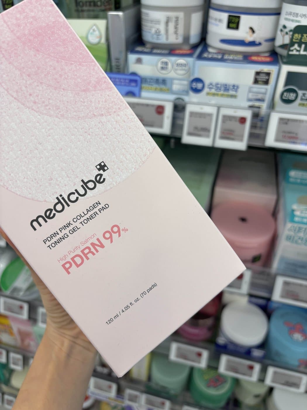 Hand holding a Medicube PDRN Pink Collagen Toning Gel Toner Pad box in a Korean beauty store, with "PDRN 99%" printed on the front
