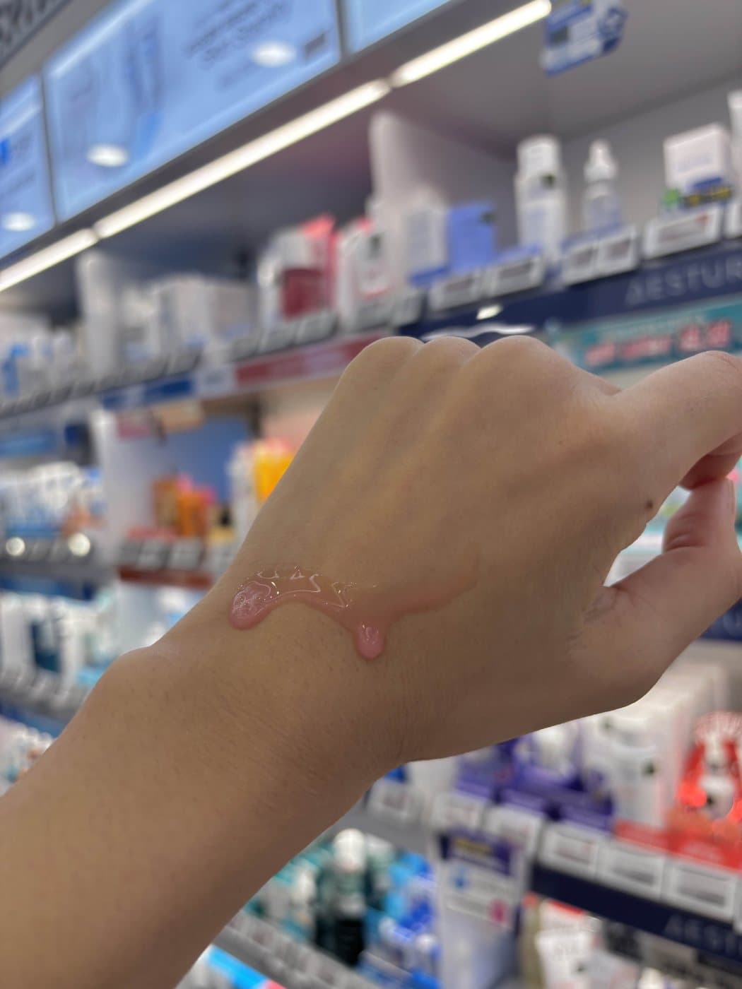 A clear pink PDRN serum swatched on the back of a hand in a Korean beauty store aisle