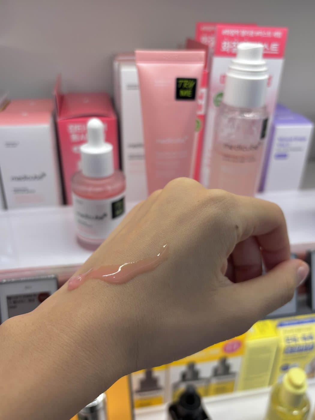Clear pink PDRN serum swatched across the back of a hand in a Korean beauty store, with a Medicube PDRN serum bottle, tube and mist on the shelf behind