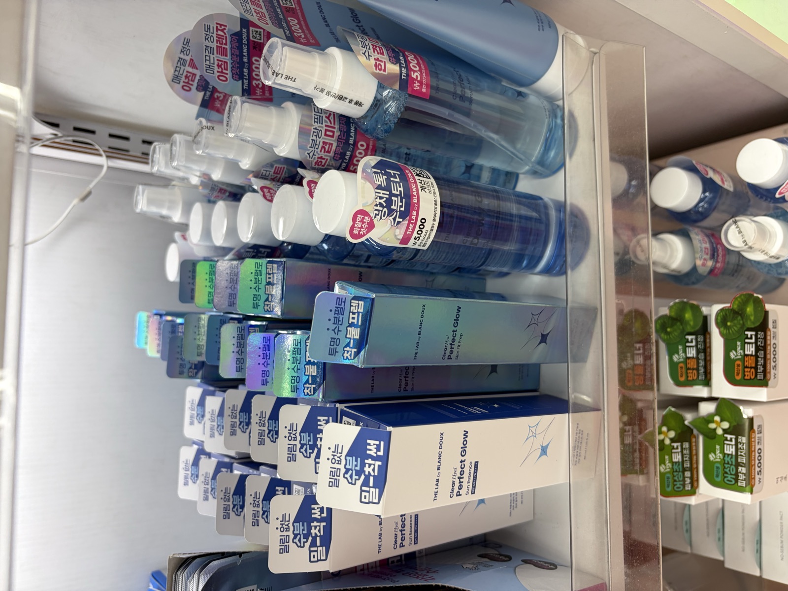 Close-up of The Lab by Blanc Doux Clear Hyal Perfect Glow Skin Fit Prep holographic boxes stacked on a Daiso shelf with Korean lettering reading chak-buteu prime cream on the spine
