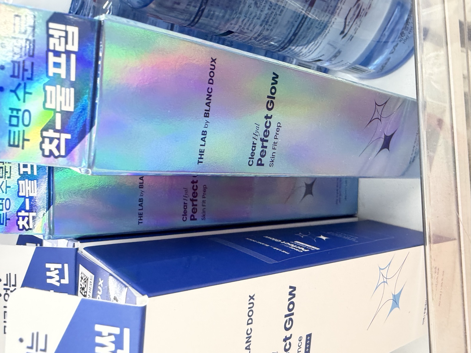 Daiso shelf display of The Lab by Blanc Doux Clear Hyal Perfect Glow Skin Fit Prep holographic boxes stacked above the blue Sun Essence box with toner bottles in the background