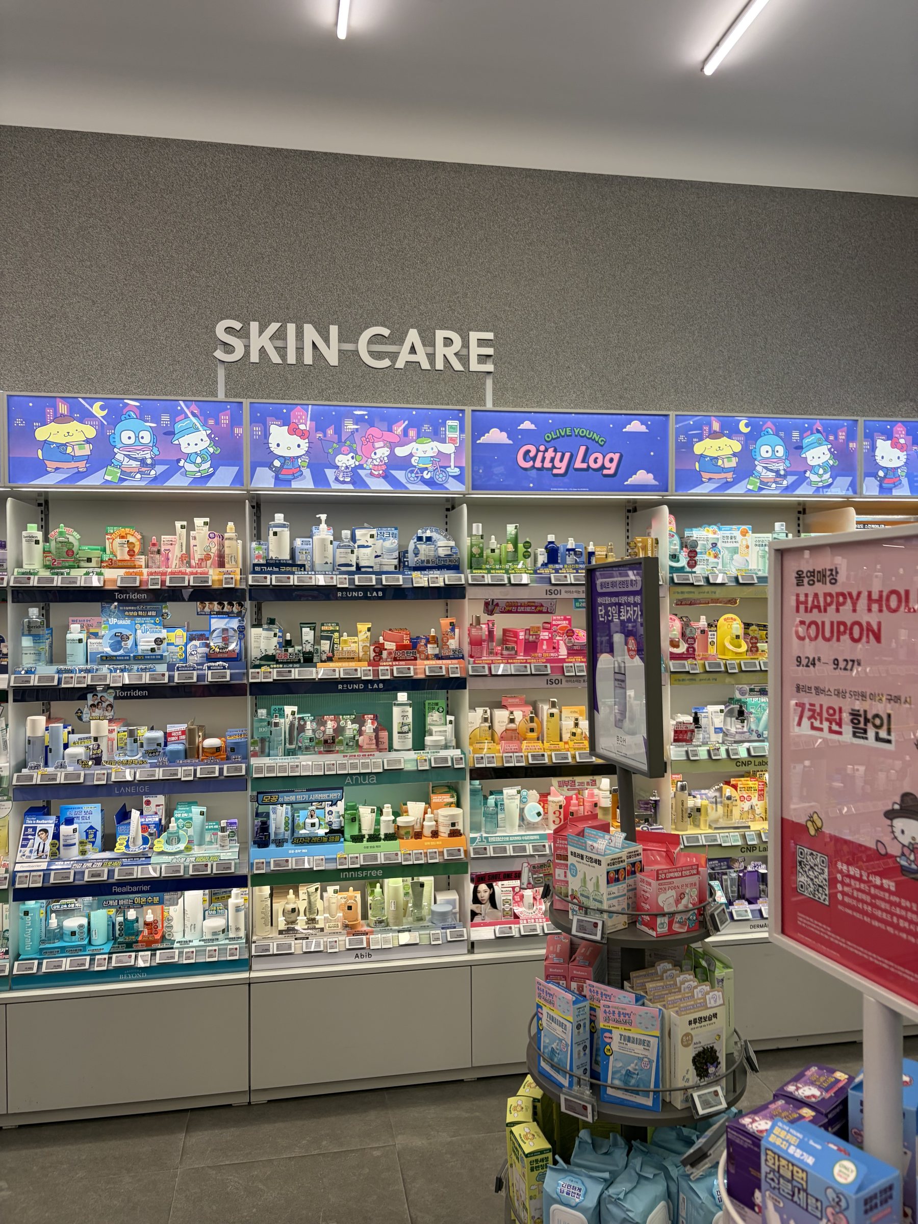 Olive Young skincare section with Sanrio City Log banners and a Happy Holiday coupon sign on the right