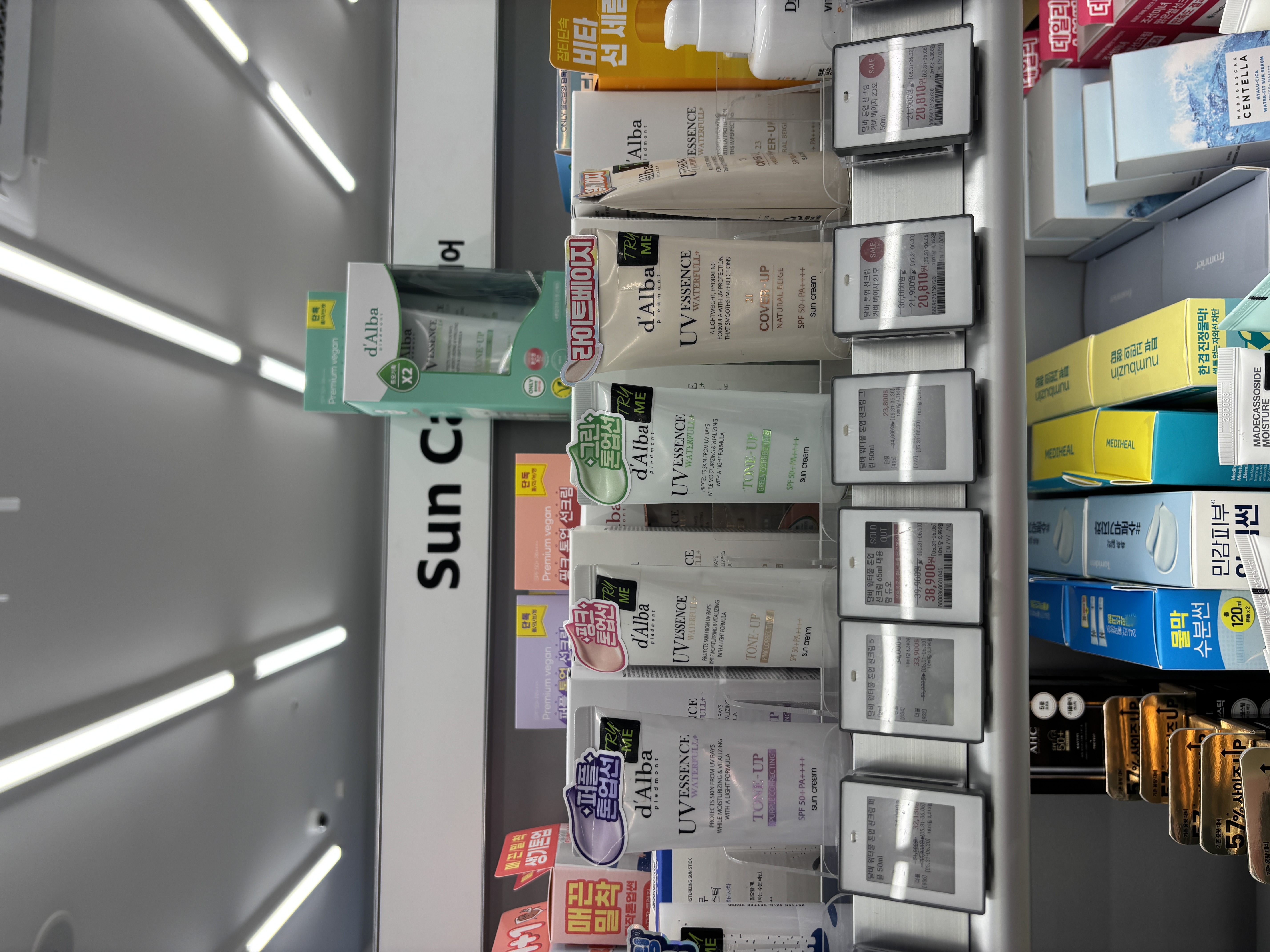 d'Alba UV Essence Waterfull sunscreen lineup at Olive Young showing Cover-Up, Tone-Up, and regular Sun Cream variants with price tags and promotional stickers